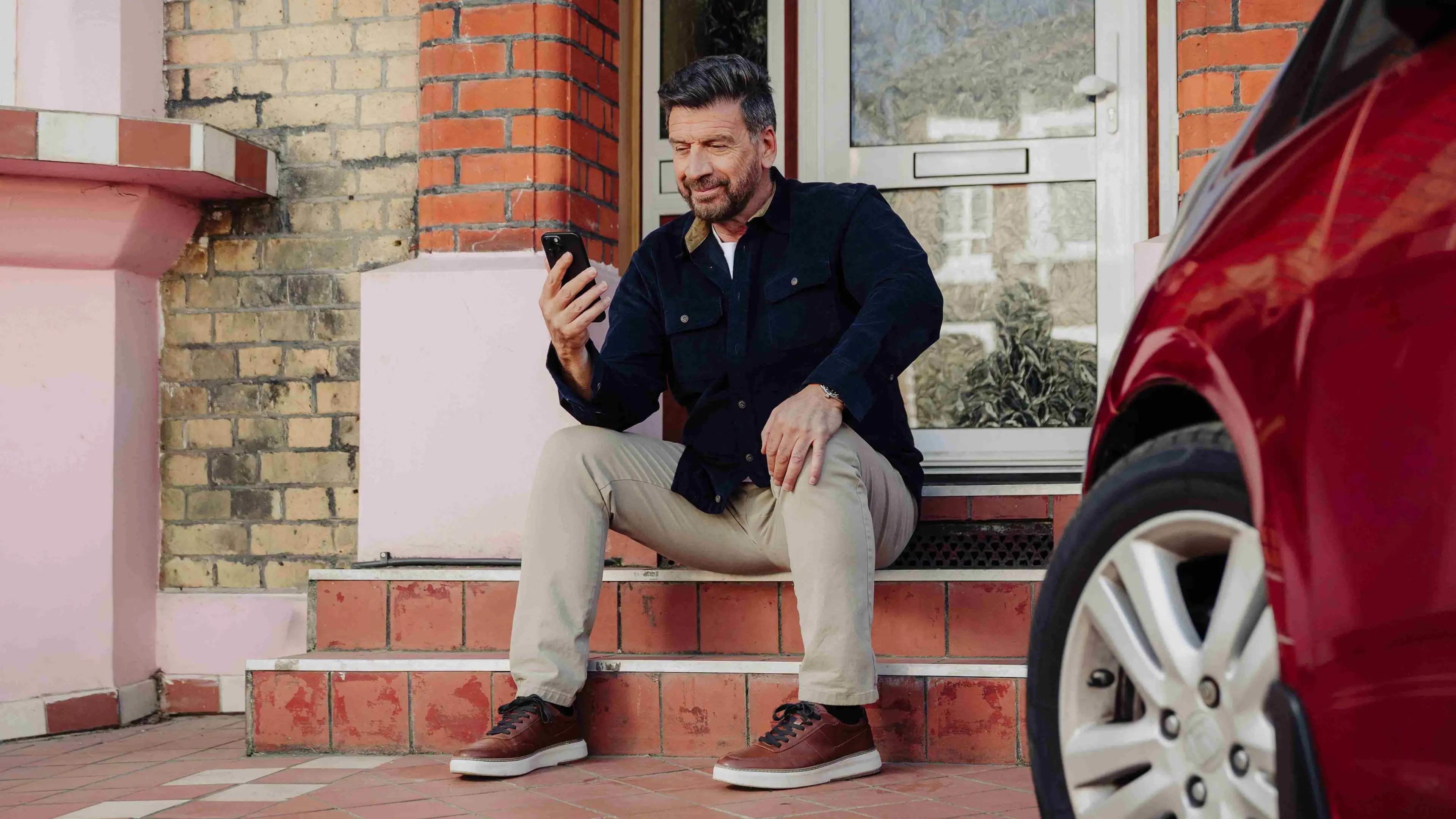 Nick Knowles seated on a doorstep looking at his phone beside a red car